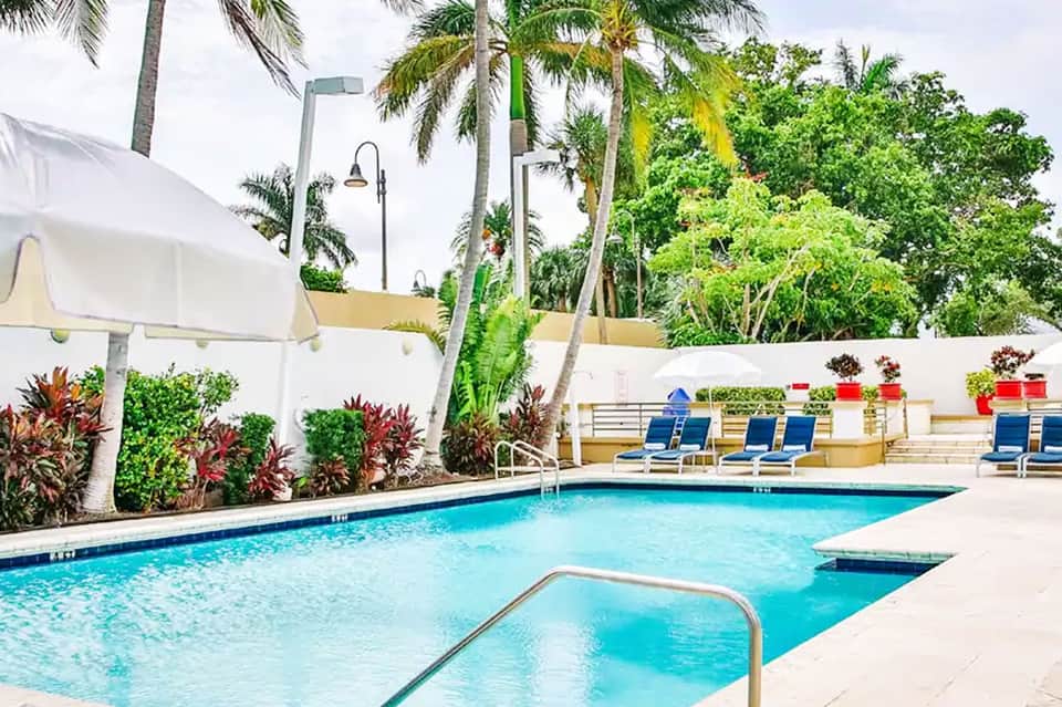 Resort pool area surrounded by white walls, palm trees, lounge chairs, and colorful tropical flowers