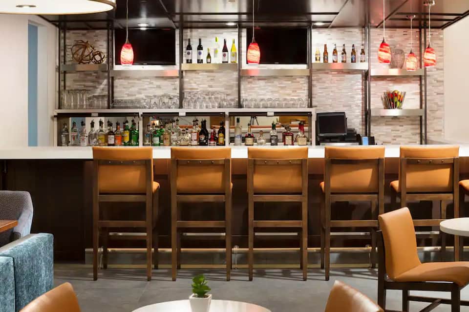 Modern bar counter with wooden stools, hanging pendant lights, and shelves of liquor bottles