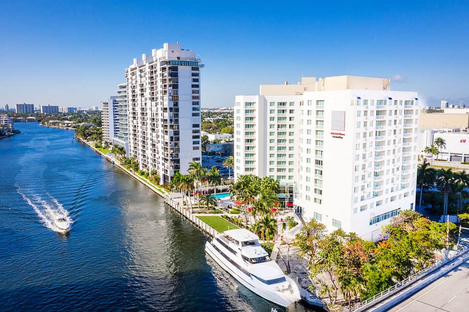 Waterfront resort with white apartment buildings, boats, palm trees, and blue waterway under clear sky
