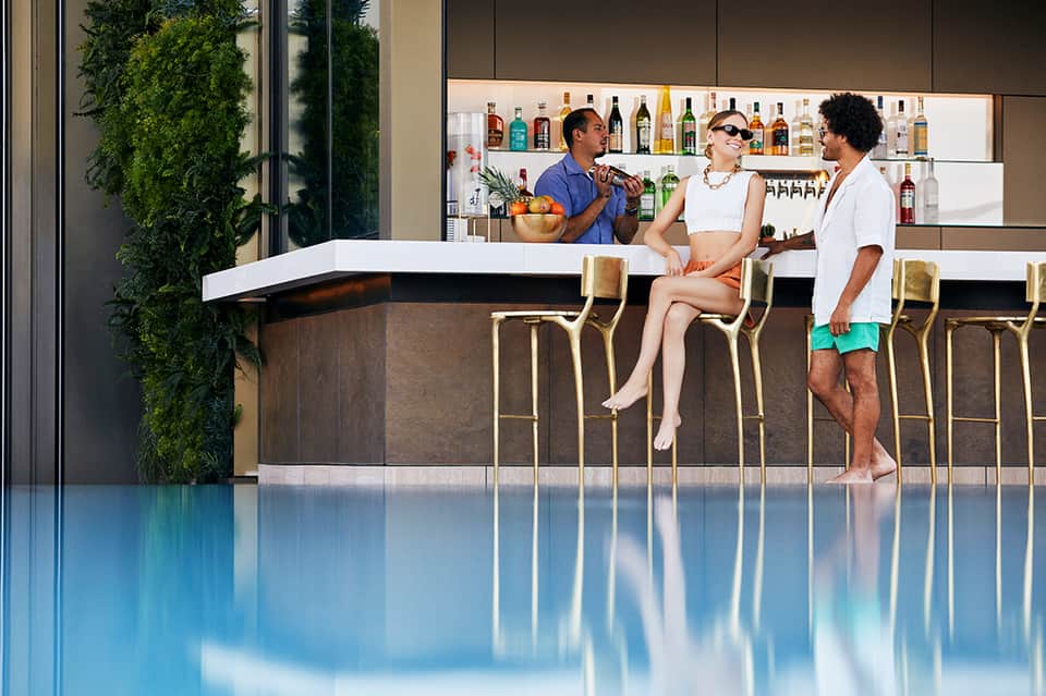 Poolside bar with three people sitting at curved white counter, backlit liquor shelves, and blue water in foreground