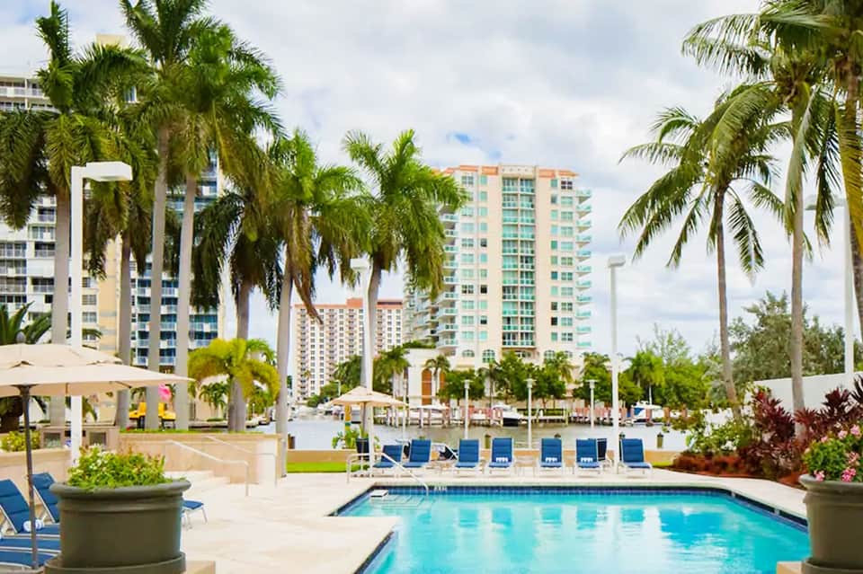 Luxury resort pool with palm trees, blue lounge chairs, and waterfront high-rise buildings in background