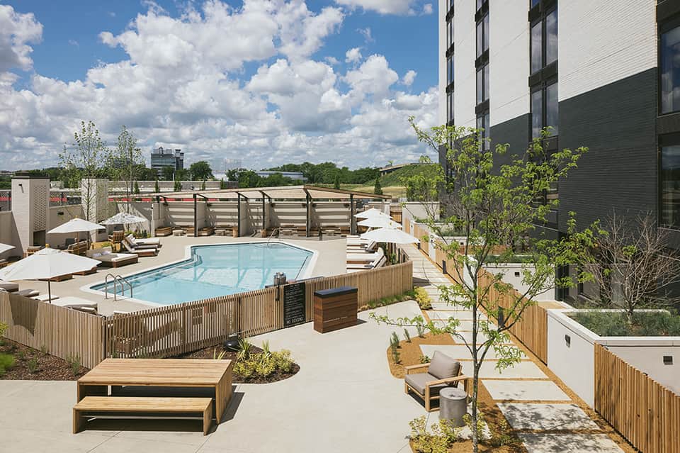 Modern rooftop pool deck with lounge chairs, umbrellas, and wooden fencing overlooking landscaped grounds