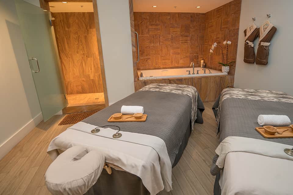 Spa treatment room with two massage beds, wooden accents, soaking tub, and plush towels