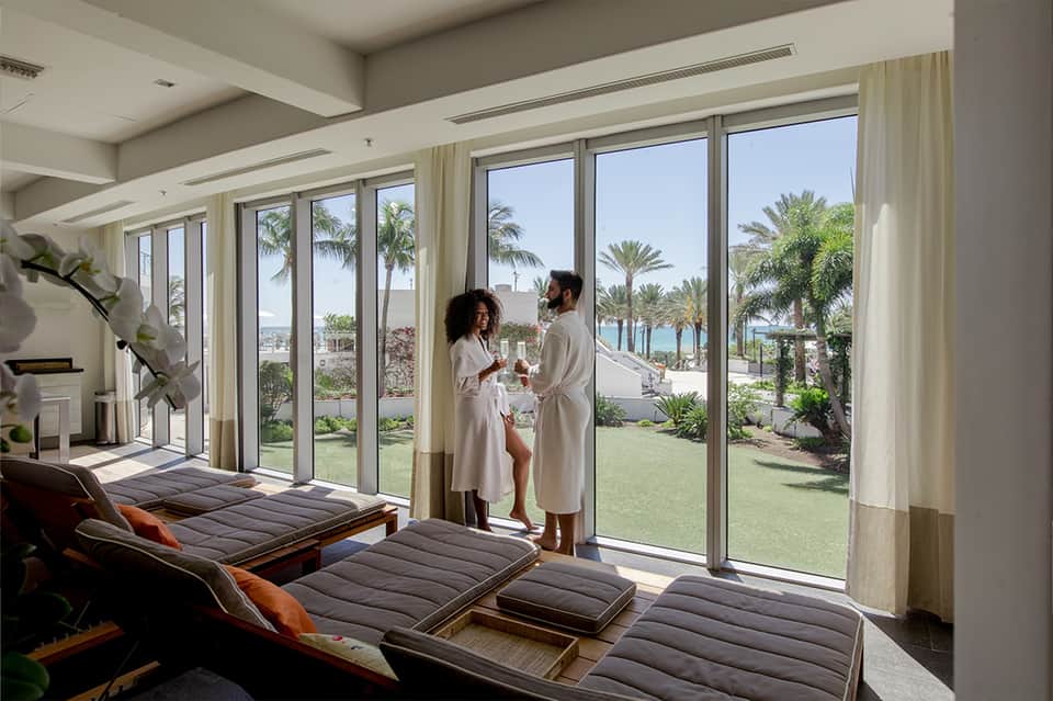 Couple in robes standing on terrace overlooking landscaped grounds with palm trees and ocean view
