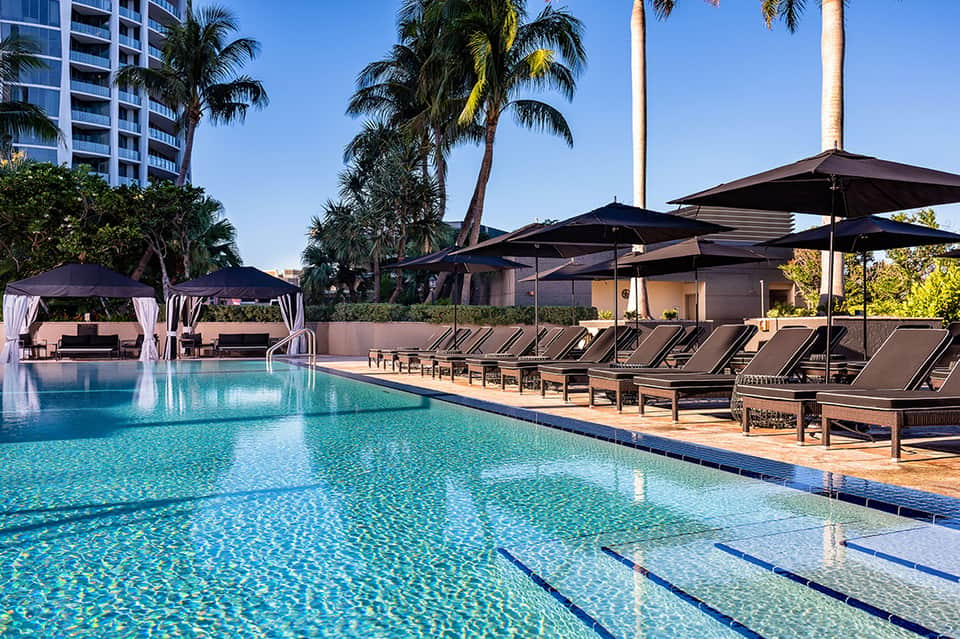 Resort pool with lane markers, lounge chairs, and dark umbrellas under palm trees and blue sky