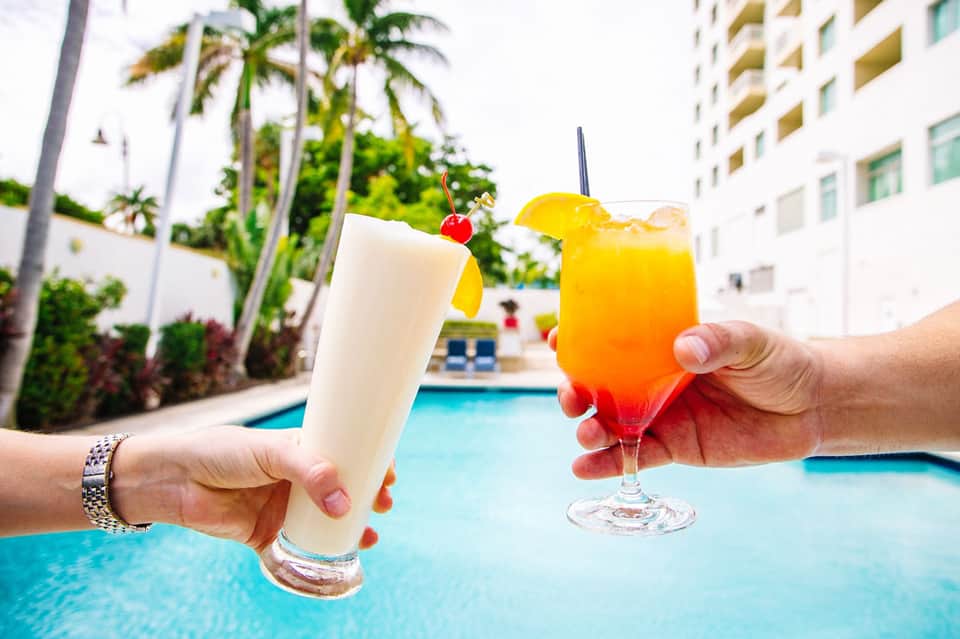 Two tropical cocktails held by pool with palm trees and resort buildings in background
