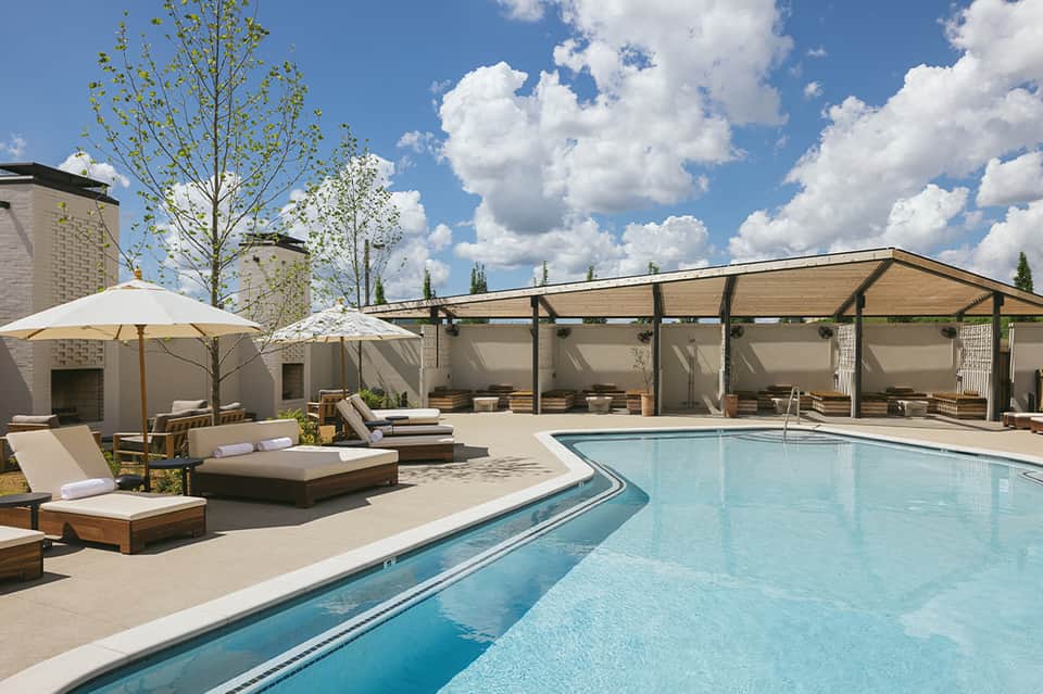 Outdoor pool deck with loungers, umbrellas, pergola, and landscaping under blue sky