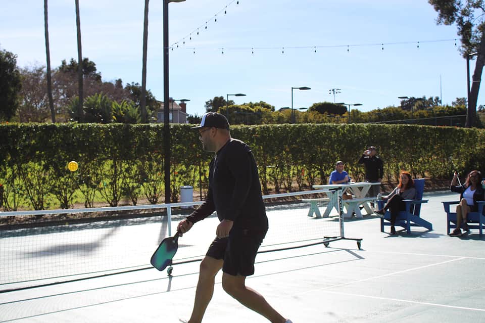 Man playing pickleball on outdoor court with vineyard views and spectators in background