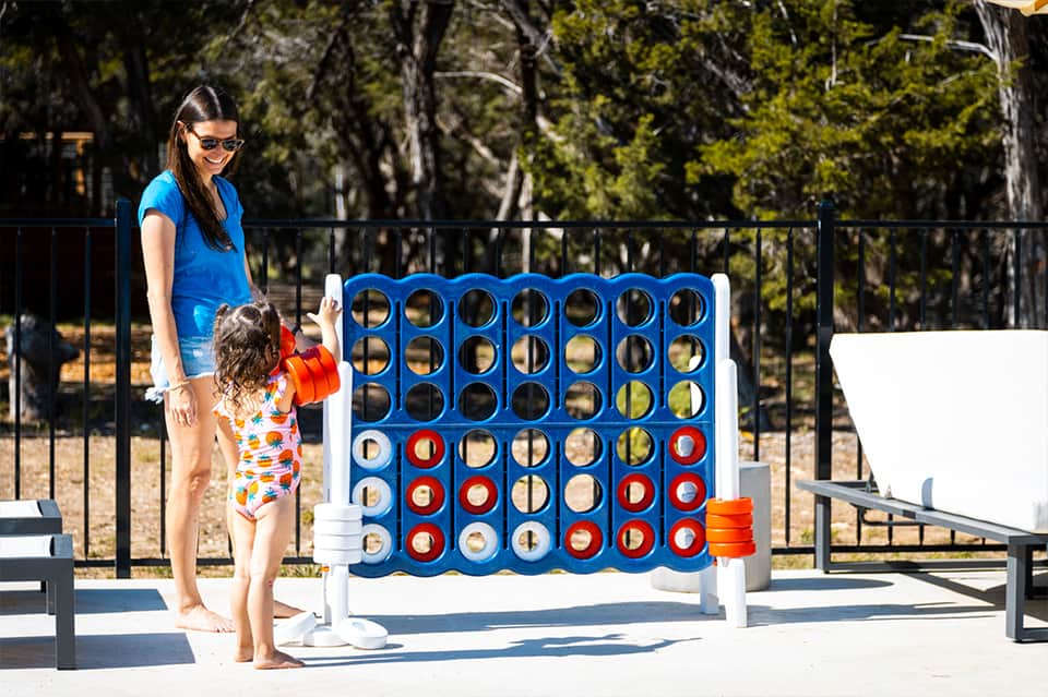 Woman and child playing oversized Connect Four game at a sunny outdoor park area with trees