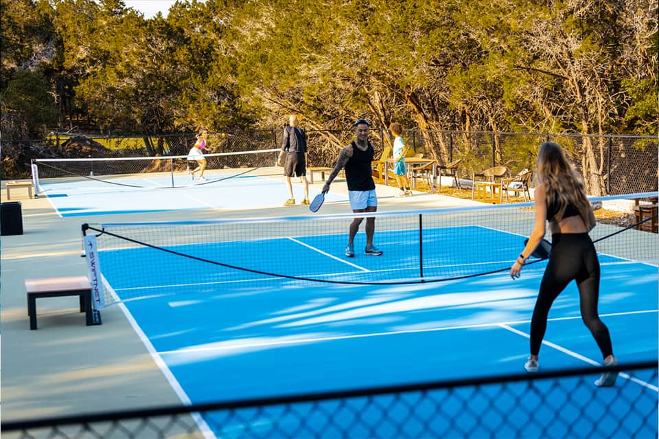 Outdoor pickleball courts with blue and white playing surfaces surrounded by trees