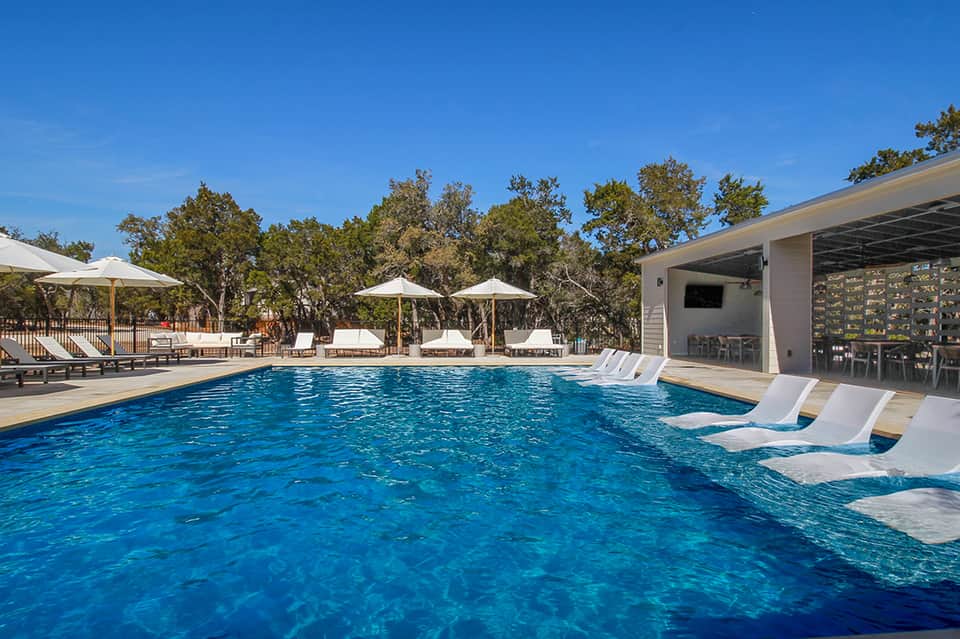 Resort swimming pool with lounge chairs, umbrellas, and modern poolside building surrounded by trees