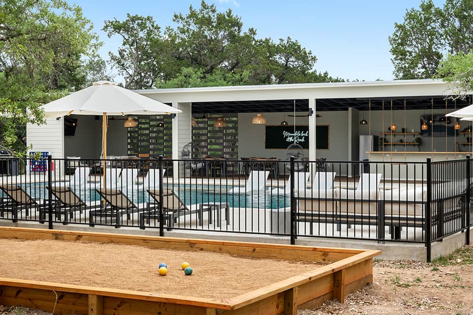 Modern poolside bar and grill with black metal fencing, white umbrellas, and bocce ball court on sandy ground