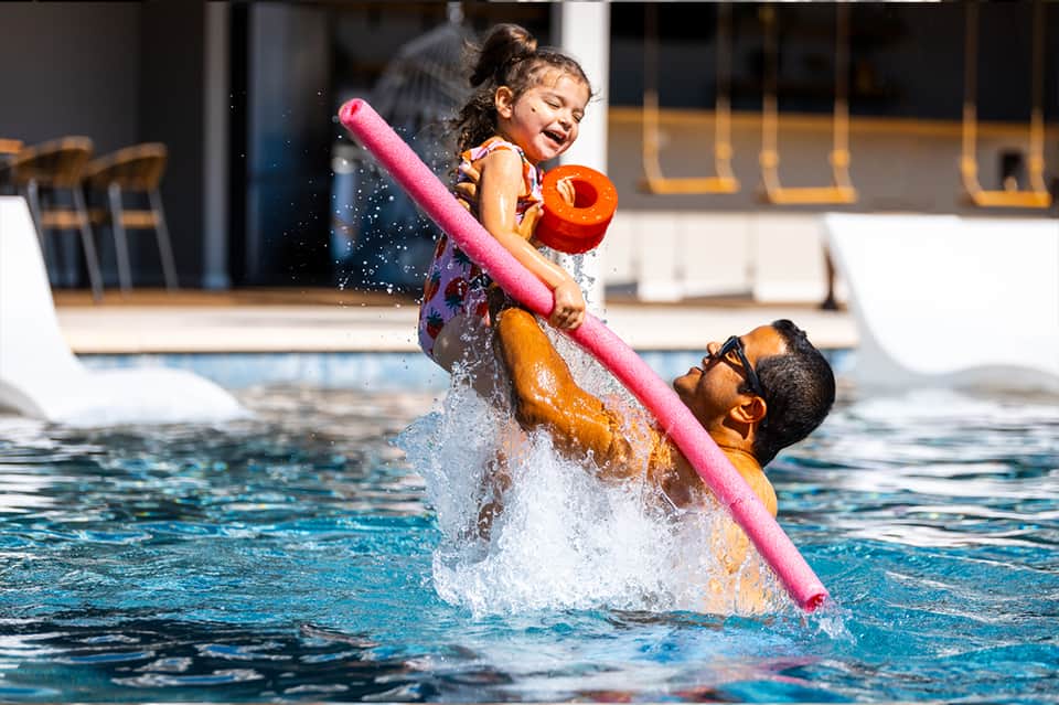 Adult lifting child in pool while playing with colorful float toys on sunny day