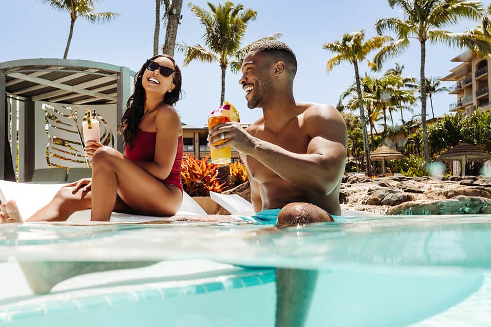Couple enjoying drinks poolside at tropical resort with palm trees and hotel buildings in background