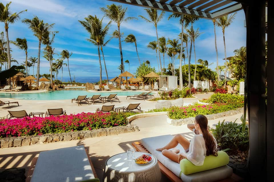 Tropical resort pool area with palm trees, lounge chairs, flowering plants, and ocean views