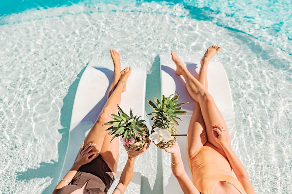 Two people on paddleboards in turquoise ocean water holding fresh pineapples