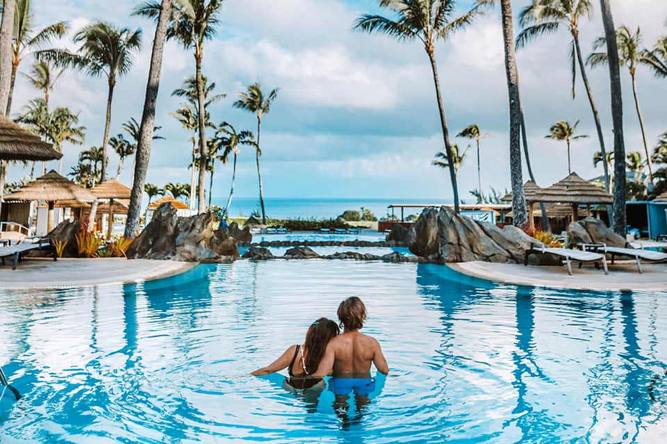 Two children wading in tropical resort pool with palm trees and ocean view in background