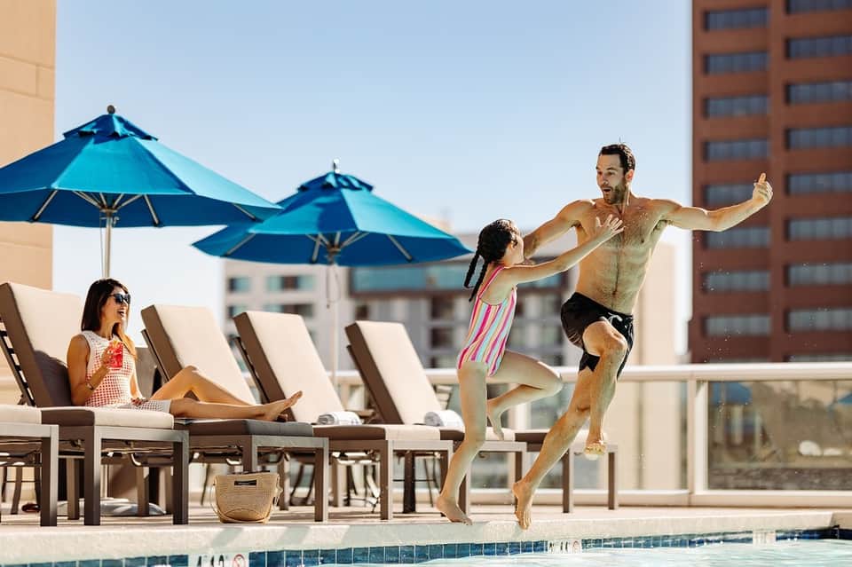Man and child jumping into pool while woman relaxes on lounge chair under blue umbrellas