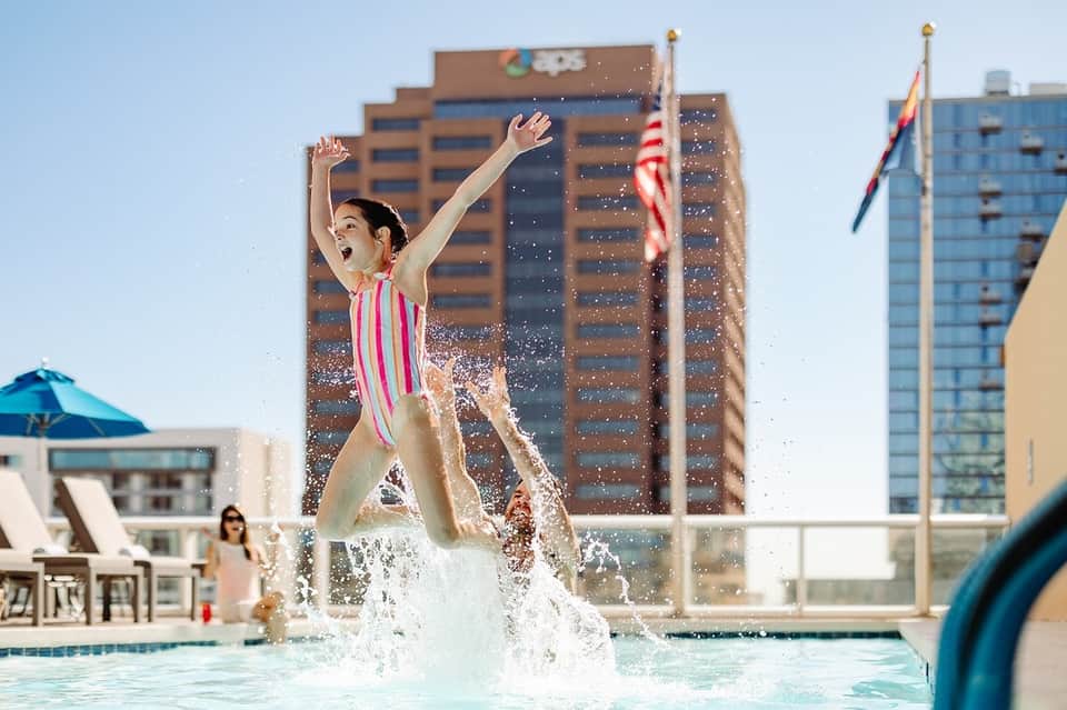 Young girl jumping in hotel pool with arms raised, high-rise buildings and urban skyline in background