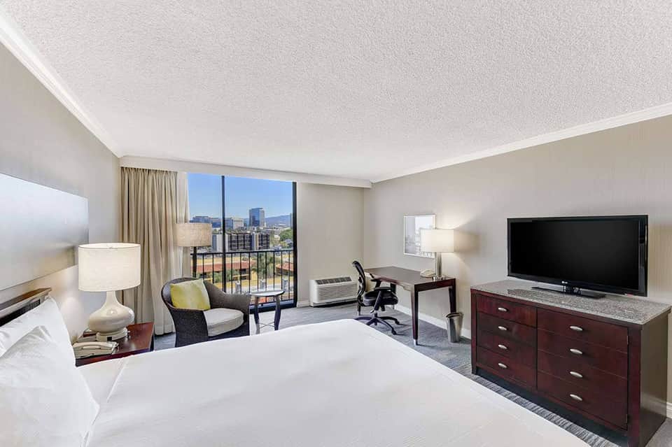Modern hotel room with king bed, work desk, TV, and balcony overlooking city