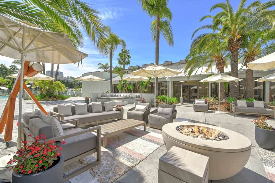 Daytime resort terrace with lounge seating, umbrellas, fire pit, and palm trees