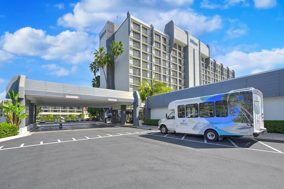 Modern hotel exterior with multi-story residential tower, covered entrance, and shuttle bus under clear blue sky
