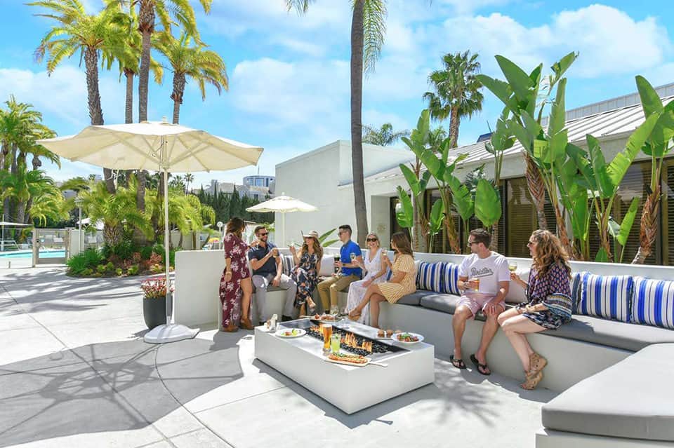 Modern poolside lounge area with white umbrellas, palm trees, and guests socializing on contemporary seating