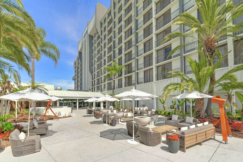 Contemporary hotel courtyard with palm trees, white umbrellas, lounge seating, and multi-story building