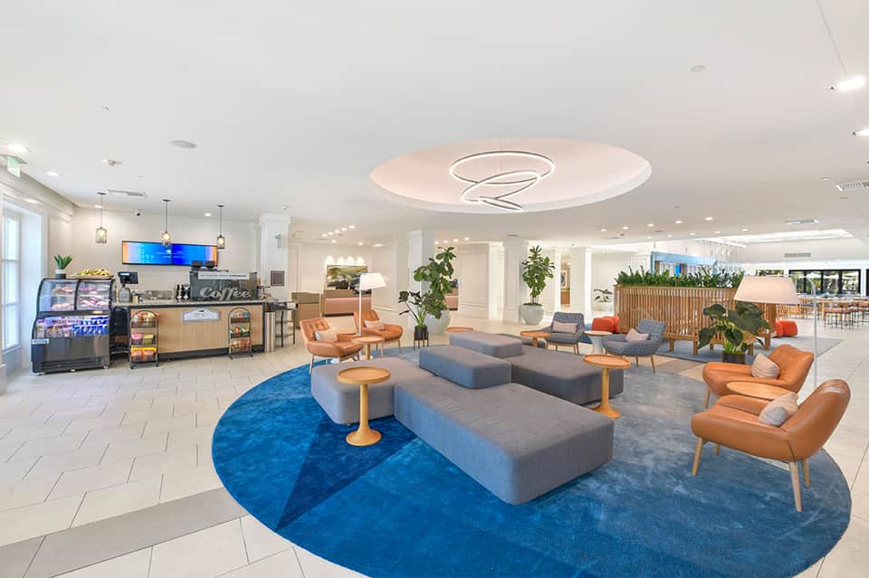 Modern hotel lobby with blue circular rug, gray seating, orange accents, coffee bar, and indoor plants