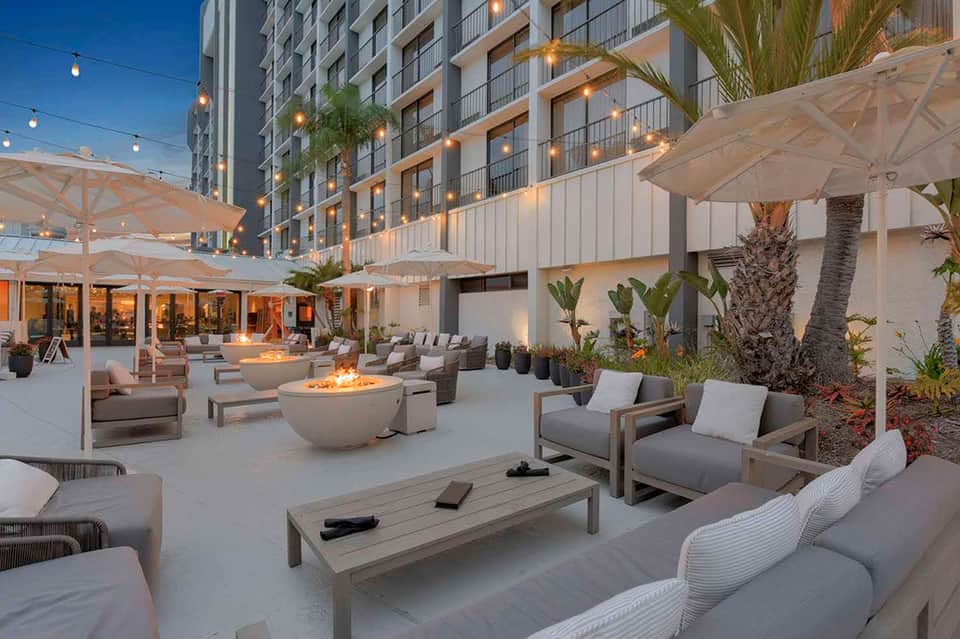 Modern resort courtyard at dusk with lounge seating, fire pits, umbrellas, and illuminated high-rise building