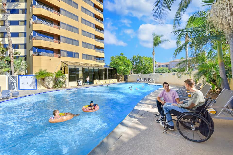Resort pool with families swimming, palm trees, and multi-story yellow building in background
