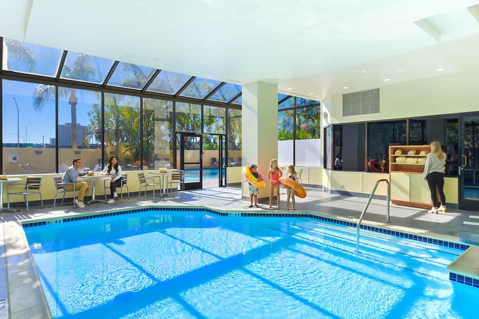 Indoor pool facility with large windows, palm trees visible outside, families enjoying swimming area