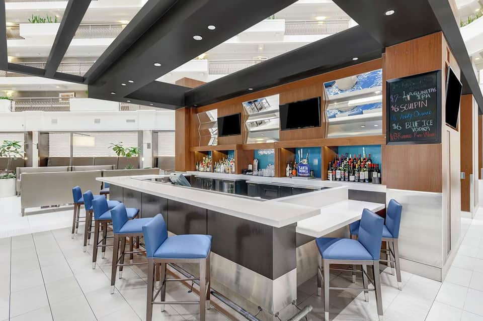 Modern hotel bar with blue bar stools, white countertop, wooden back panel with shelving, and black ceiling beams.