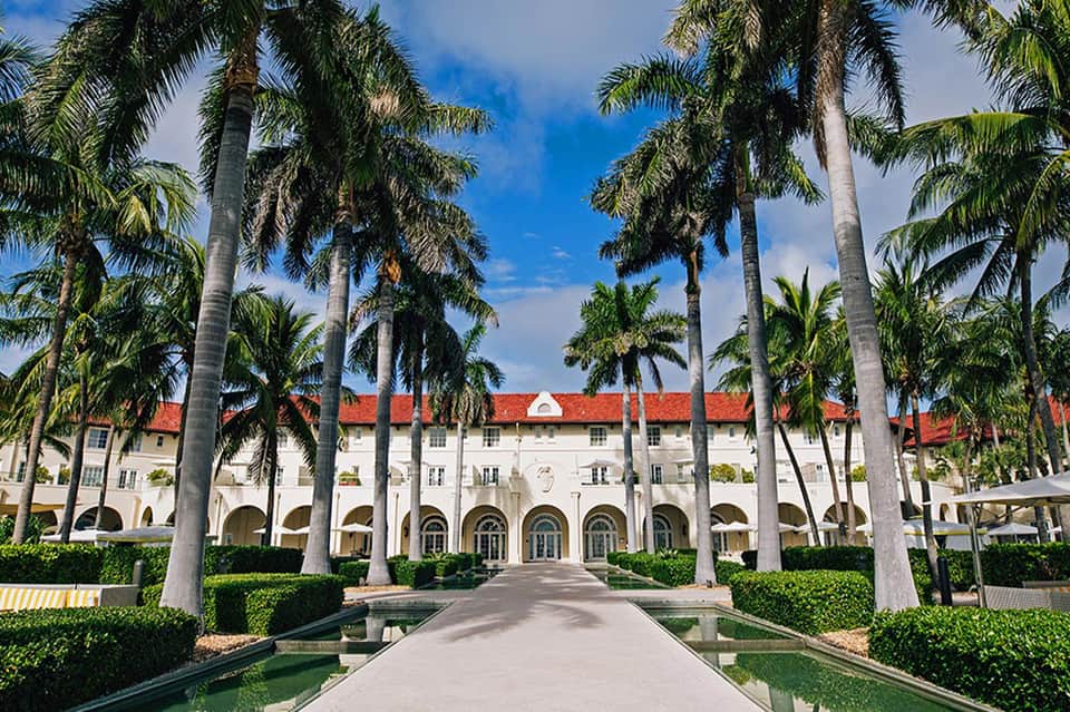 Historic white colonial building with red roof framed by tall palm trees and manicured hedges