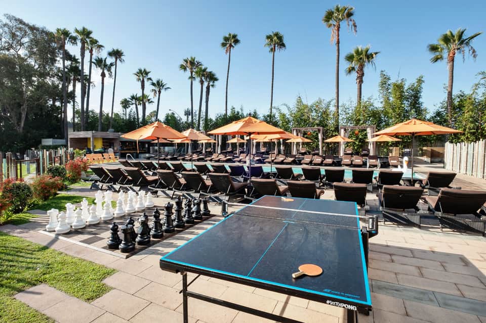 Outdoor recreational area with ping pong table, giant chess set, lounge chairs, orange umbrellas, and palm trees