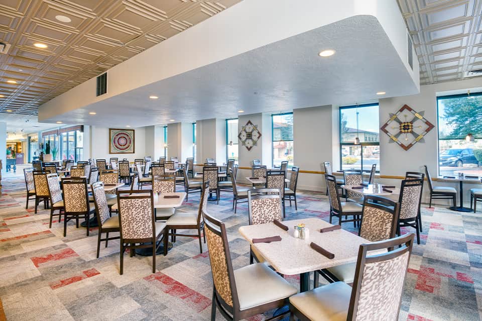 Bright dining area with patterned chairs, tables, decorative wall art, and large windows overlooking parking lot