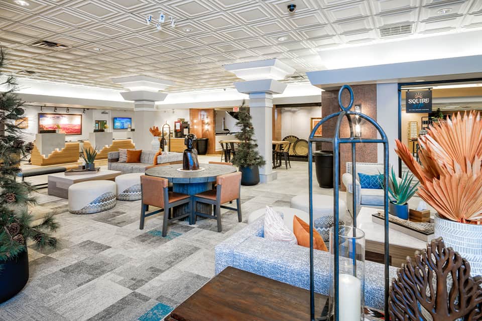 Modern hotel lobby with blue and orange accents, geometric ceiling, comfortable seating areas, and tropical plants