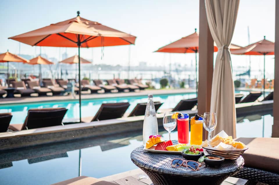 Poolside dining table with beverages, appetizers, and ocean city skyline view under orange umbrellas