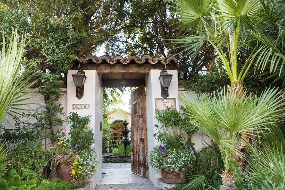 Spanish colonial-style white stucco entrance with ornate arch, weathered wooden door, and vibrant flowering plants