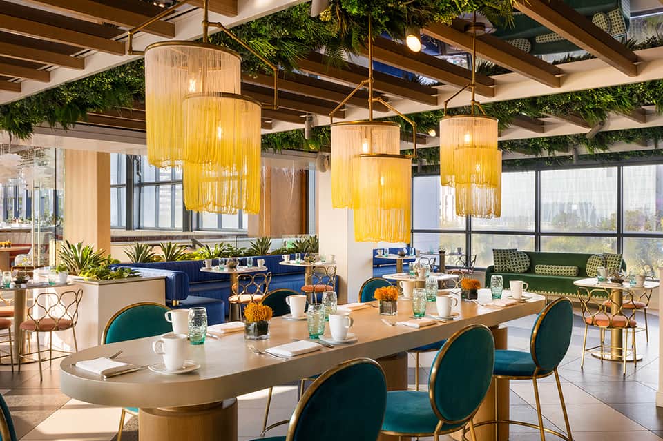 Modern meeting space with wooden ceiling beams, yellow pendant lights, green plants, blue seating, and large windows
