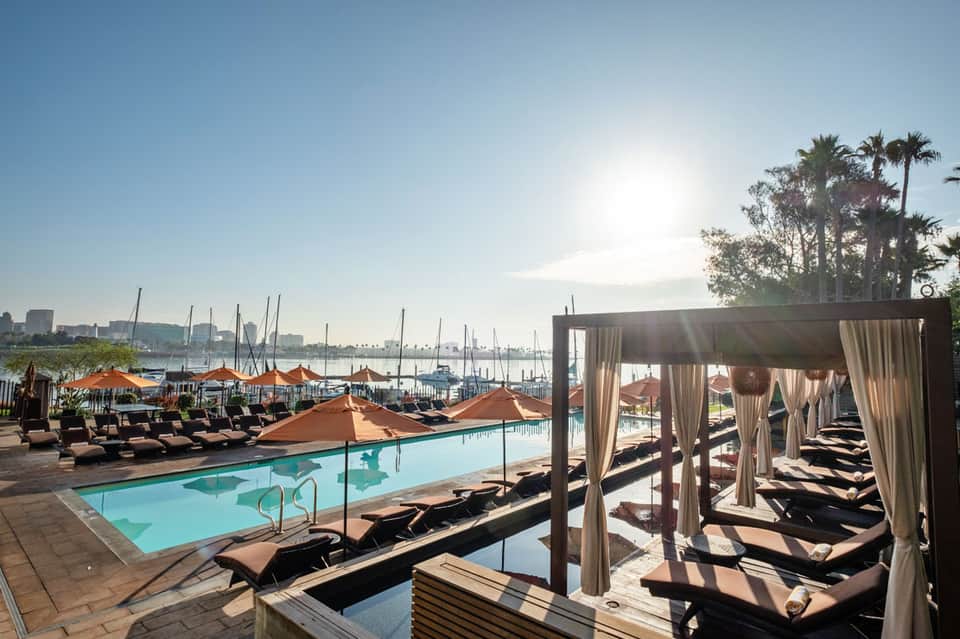 Luxury resort pool with orange umbrellas, lounge chairs, and cabanas overlooking marina with sailboats