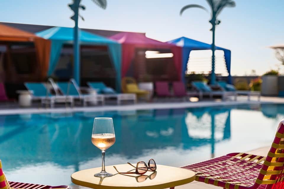 Poolside wine glass and sunglasses on table with colorful cabanas and palm trees in background