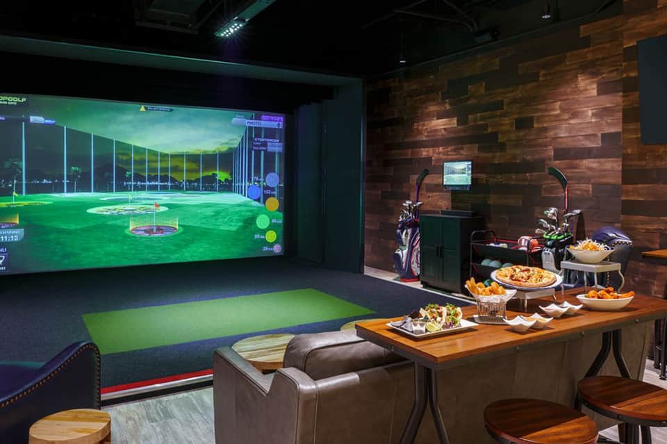 Indoor golf simulator lounge with bar seating, snacks, and wood-paneled accent wall
