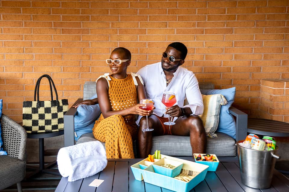 Couple sitting on a brick patio enjoying cocktails with snacks and a beach bag nearby