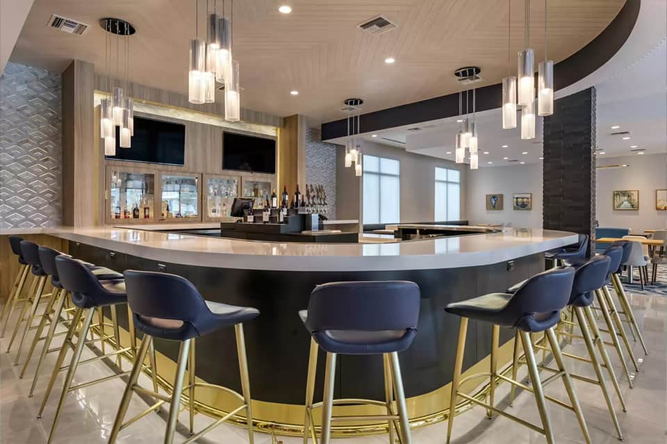 Contemporary hotel bar with curved black countertop, blue bar stools, pendant lighting, and wood finishes