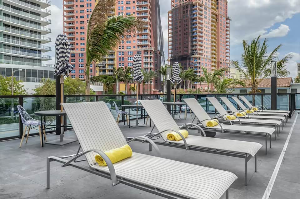Rooftop lounge with white loungers, yellow pillows, palm trees, and city skyline view