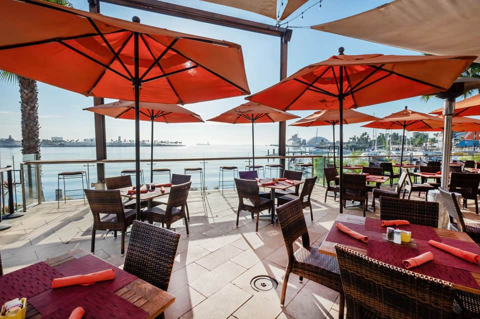 Waterfront dining terrace with red umbrellas, wicker chairs, and bay views