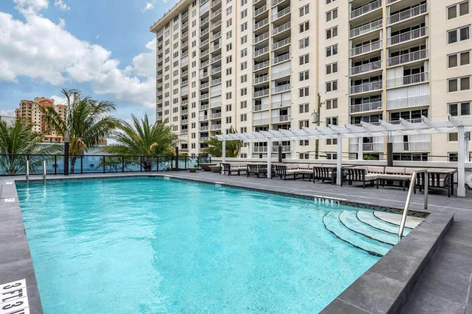 Resort pool surrounded by multi-story hotel buildings with palm trees and lounge seating