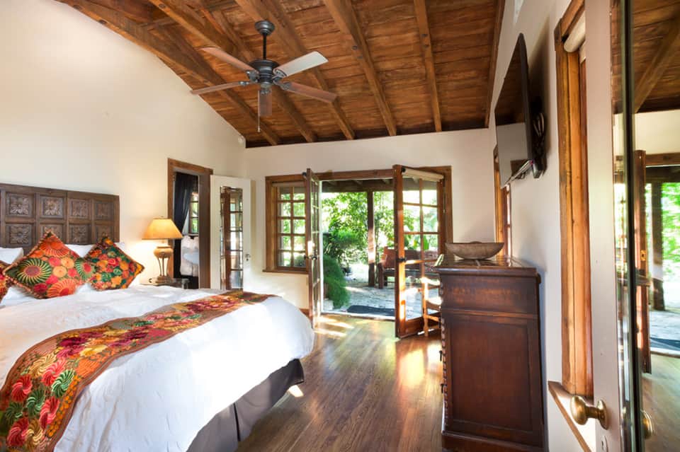Luxury bedroom with wooden beamed ceiling, colorful pillows, wooden furniture, and doors opening to garden view
