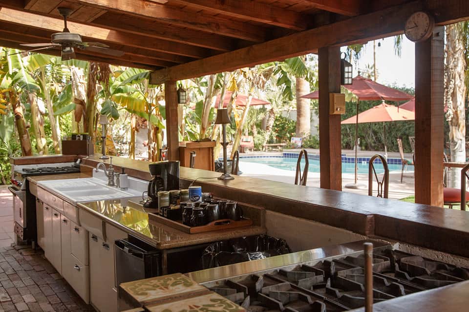 Covered outdoor kitchen with stainless steel appliances, island counters, and pool view through wooden beams